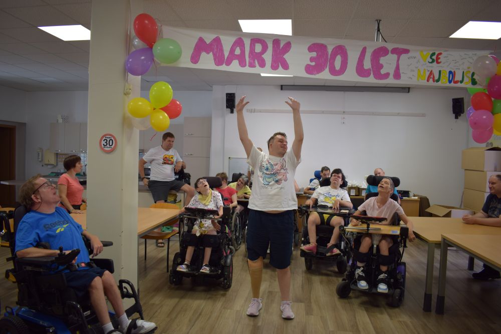 Mark stoji pod transparentom na katerem piše MARK 30 LET. Ima dvignjene roke in se smeje. V ozadju so ostali uporabniki na vozičkih.