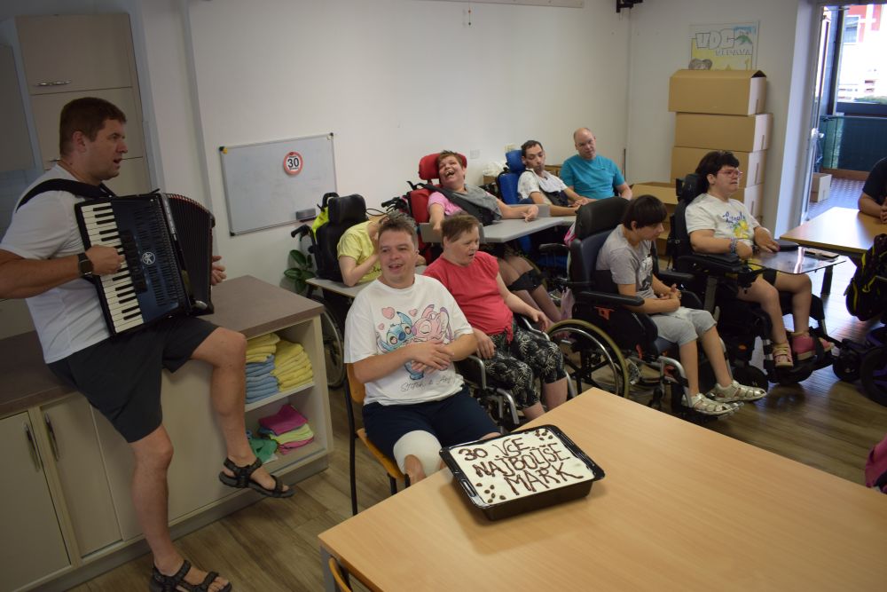 Marko igra Marku na harmoniko. V ozadju so uporabniki. Pred Markom je tiramisu na katerem piše MARK VSE NAJBOLJŠE.
