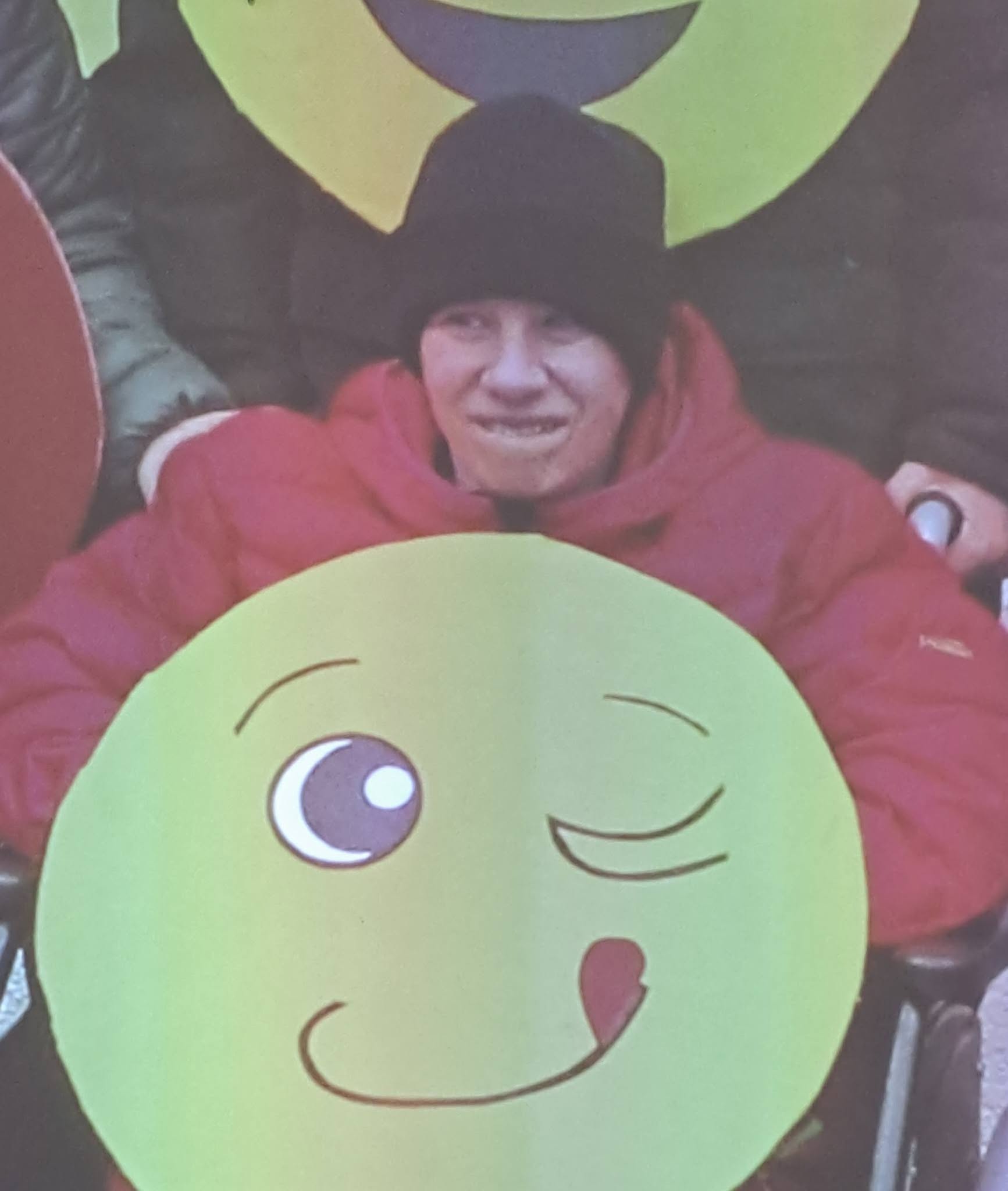 Na fotografiji je Patricija Šušmelj, v rdeči jakni in v rokah drži rumenega smeškota oz. sonce. Fotografija je nastala na enem od pustovanj v VDCju v enoti Ajdovščina, kamor je bila pokojna Patricija vključena pred leti.