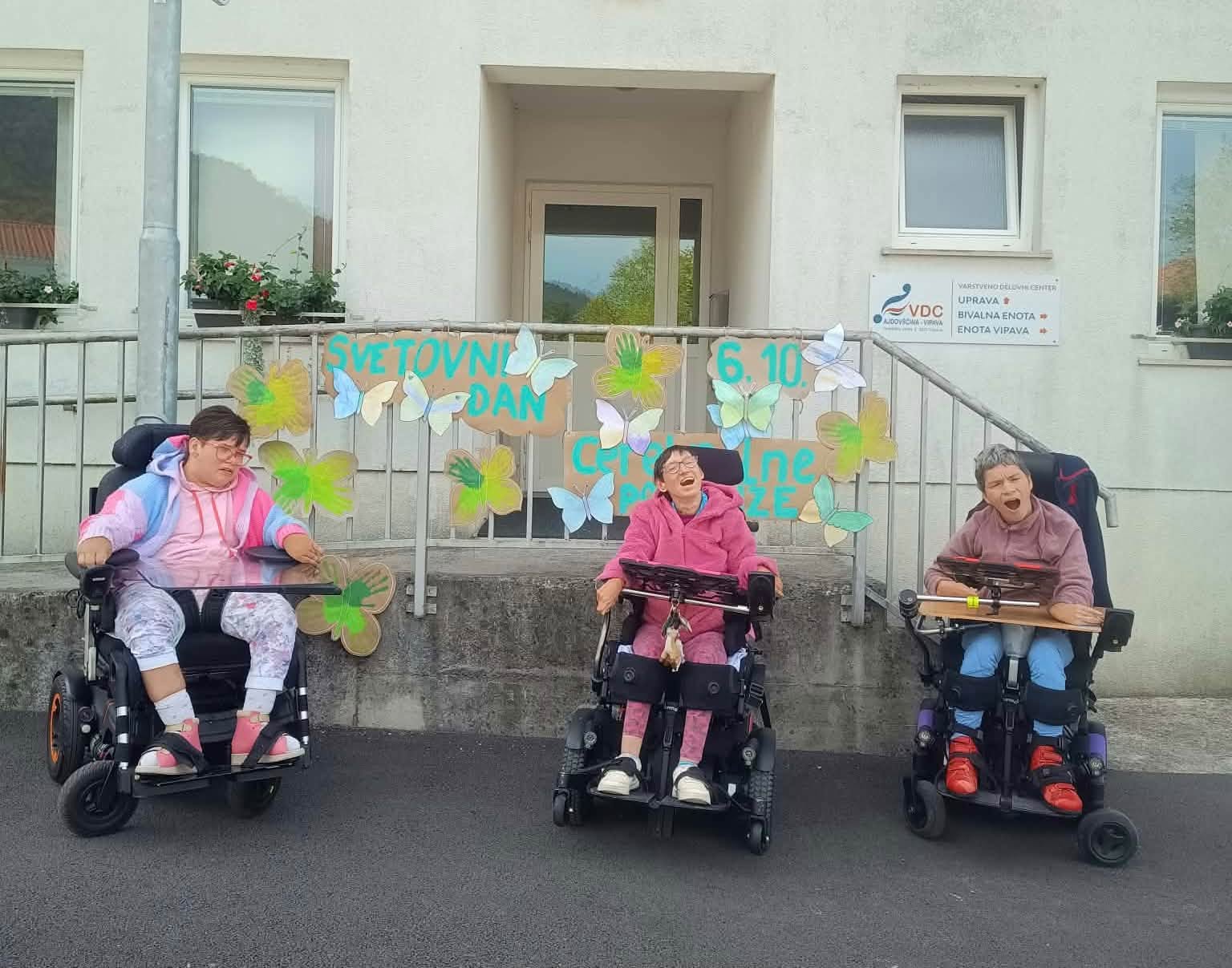 Jožica, ANdreja in Katka z vozički pred stavbo VDC v Vipavi. Postavljene so pred ograjo ob vhodu v stavbo. Na ograji je napis Svetovni dan cerebralne paralize, datum 6.10. in zeleni metulji različnih odtenkov in velikosti